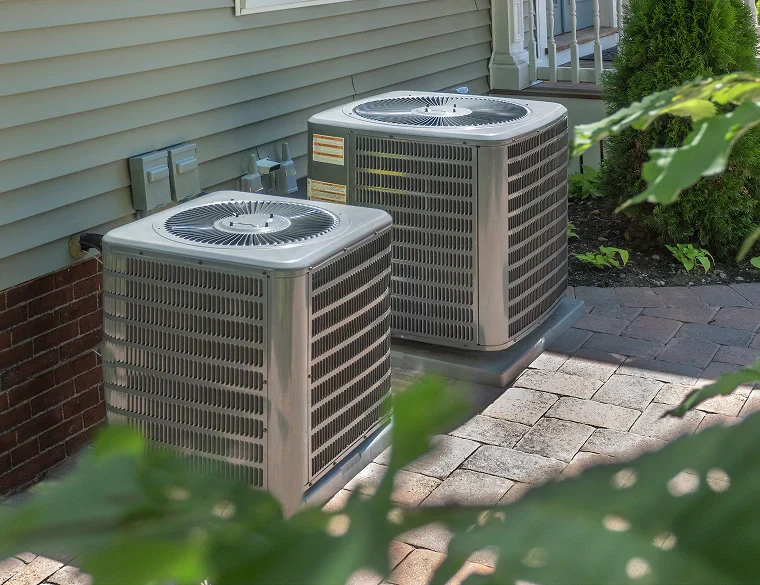 Two air conditioning units sit side by side on a stone patio beside a house, partially obscured by green foliage