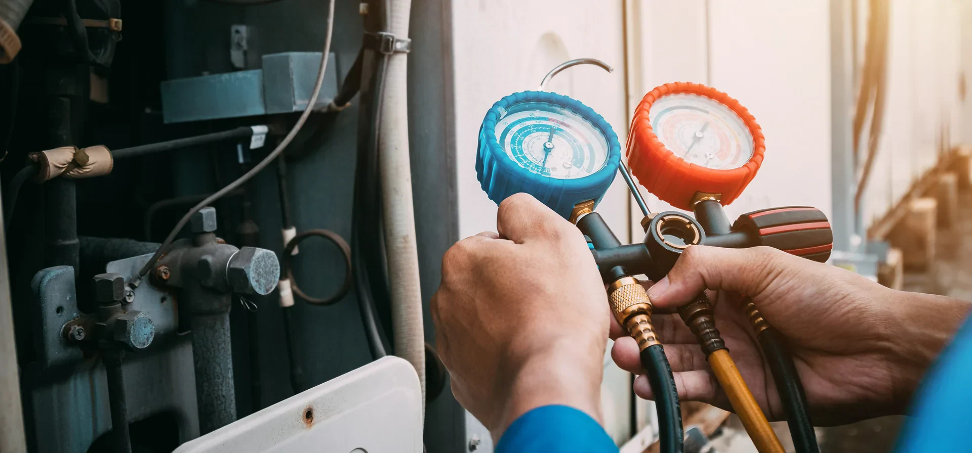 A technician holds blue and red pressure gauges while working on an air conditioning unit, with various pipes and tools in the background