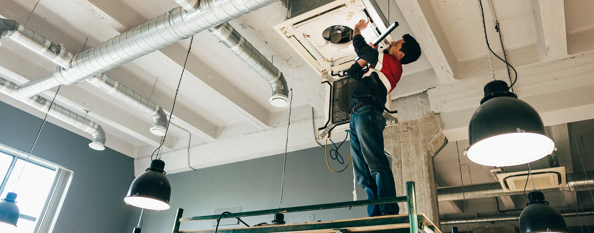 A worker on a scaffold repairs an air conditioning unit, surrounded by industrial pipes and hanging lights in a modern interior space